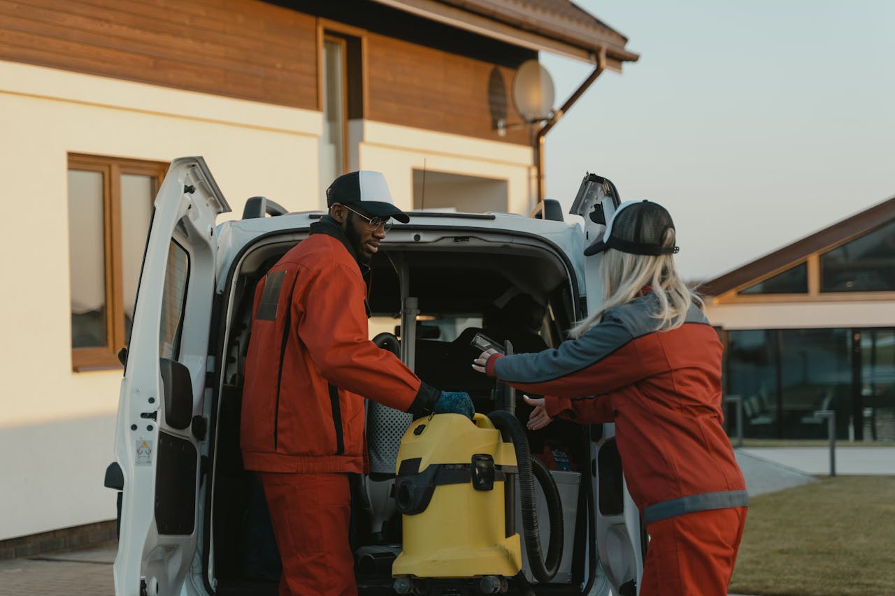 Two cleaners in red uniforms load a vacuum into a white van outdoors.